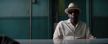 Movie still from “2 Guns” (2013), directed by Baltasar Kormákur – A man wearing a hat sitting at a table in front of a green door; Close Up shot, Low angle