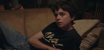 Movie still from “20th Century Women” (2016), directed by Mike Mills – A young man sitting on a couch looking at the camera; Medium shot, High angle
