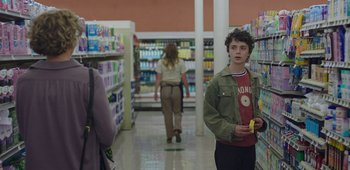 Movie still from “20th Century Women” (2016), directed by Mike Mills – A boy is holding a banana in a store; Medium shot, Over the shoulder angle
