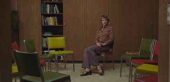 Movie still from “20th Century Women” (2016), directed by Mike Mills – A woman sitting on a chair in front of a bookshelf; Wide shot, Low angle