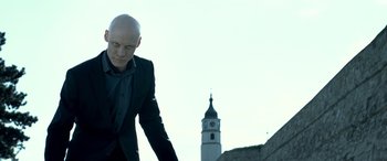 Movie still from “3 Days to Kill” (2014), directed by McG – A man in a suit and tie standing in front of a clock tower; Medium shot, Low angle