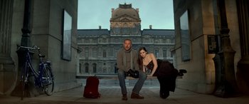 Movie still from “3 Days to Kill” (2014), directed by McG – A man and a woman posing for a picture in front of a building; Wide shot, Low angle