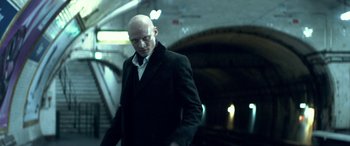 Movie still from “3 Days to Kill” (2014), directed by McG – A bald man in a black suit standing next to a tunnel; Medium shot, Low angle