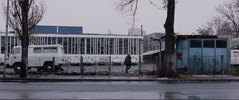 Movie still from “4 Months, 3 Weeks and 2 Days” (2007), directed by Cristian Mungiu – A person walking on a sidewalk near a parking lot; Extreme Wide shot, Low angle