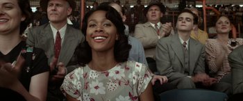 Movie still from “42” (2013), directed by Brian Helgeland – A woman smiles while sitting in a crowd; Medium shot, Over the shoulder angle
