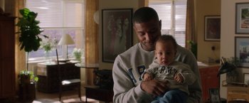 Movie still from “A Journal for Jordan” (2021), directed by Denzel Washington – A man holding a baby in his arms in a living room; Medium shot, Over the shoulder angle