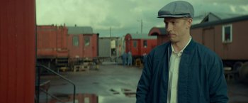 Movie still from “A Man Called Ove” (2015), directed by Hannes Holm – A man wearing a hat standing in front of a train yard; Medium shot, Over the shoulder angle