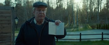 Movie still from “A Man Called Ove” (2015), directed by Hannes Holm – An older man holding a book in front of a forest; Close Up shot, Over the shoulder angle