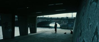 Movie still from “A Most Wanted Man” (2014), directed by Anton Corbijn – A man standing under a bridge near a body of water; Wide shot, Over the shoulder angle