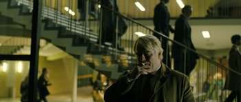 Movie still from “A Most Wanted Man” (2014), directed by Anton Corbijn – A man smoking a cigarette in front of a staircase; Medium shot, Over the shoulder angle