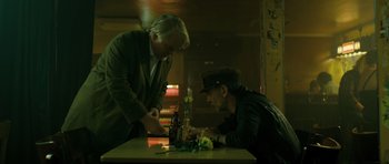Movie still from “A Most Wanted Man” (2014), directed by Anton Corbijn – A man sitting at a table next to another man holding a bottle; Medium shot, Over the shoulder angle
