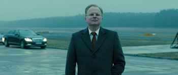 Movie still from “A Most Wanted Man” (2014), directed by Anton Corbijn – A man wearing a suit and tie standing in front of a body of water; Medium shot, Low angle