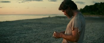 Movie still from “A Quiet Place Part II” (2020), directed by John Krasinski – A man standing on a beach holding a sandwich; Medium shot, Low angle