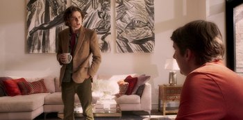 Movie still from “A Rainy Day in New York” (2019), directed by Woody Allen – A man standing in a living room holding a coffee mug; Medium shot, Over the shoulder angle