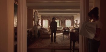 Movie still from “A Rainy Day in New York” (2019), directed by Woody Allen – A man standing in a living room in front of a fireplace; Wide shot, Low angle