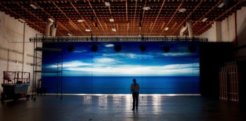 Movie still from “A Rainy Day in New York” (2019), directed by Woody Allen – A man standing in front of a large picture of the ocean; Extreme Wide shot, Low angle