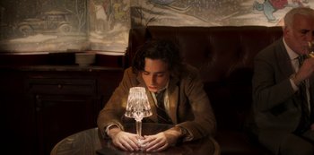 Movie still from “A Rainy Day in New York” (2019), directed by Woody Allen – A man sitting in front of a table with a wine glass on top of it; Medium shot, High angle