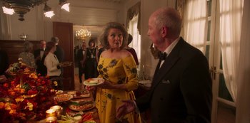 Movie still from “A Rainy Day in New York” (2019), directed by Woody Allen – An older woman in a yellow dress and an older man in a black suit stand in front of a table of food; Medium shot, Over the shoulder angle