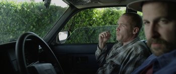 Movie still from “A Thousand Junkies” (2017), directed by Tommy Swerdlow – A man sitting in the driver's seat of a car smoking; Medium shot, Low angle