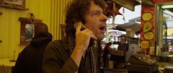 Movie still from “A Thousand Junkies” (2017), directed by Tommy Swerdlow – A man is talking on a cell phone while wearing a scarf around his neck; Close Up shot, Low angle