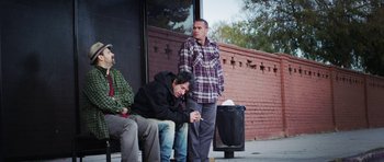 Movie still from “A Thousand Junkies” (2017), directed by Tommy Swerdlow – A group of men sitting on a bench next to a building; Wide shot, Low angle