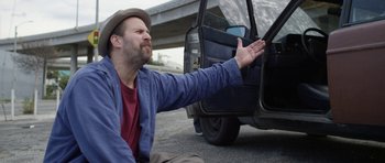 Movie still from “A Thousand Junkies” (2017), directed by Tommy Swerdlow – A man sitting on the side of the road next to a car; Medium shot, Over the shoulder angle