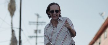 Movie still from “A Thousand Junkies” (2017), directed by Tommy Swerdlow – A man in sunglasses and a striped shirt is smiling; Medium shot, Low angle