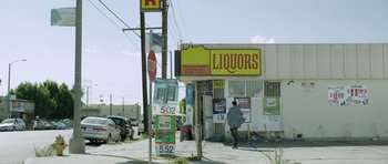 Movie still from “A Thousand Junkies” (2017), directed by Tommy Swerdlow – A man walking down the street in front of a liquor store; Extreme Wide shot, Low angle