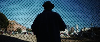 Movie still from “A Thousand Junkies” (2017), directed by Tommy Swerdlow – A man wearing a hat standing in front of a chain link fence; Medium shot, Over the shoulder angle