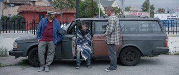 Movie still from “A Thousand Junkies” (2017), directed by Tommy Swerdlow – Three men standing in front of a parked car on the side of the road; Wide shot, Over the shoulder angle