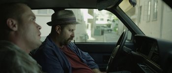 Movie still from “A Thousand Junkies” (2017), directed by Tommy Swerdlow – A man sitting in the driver's seat of a car wearing a hat; Close Up shot, Low angle