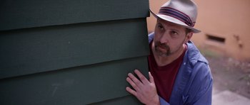 Movie still from “A Thousand Junkies” (2017), directed by Tommy Swerdlow – A man with a hat peeking over the side of a house; Close Up shot, Low angle