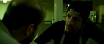 Movie still from “A Thousand Junkies” (2017), directed by Tommy Swerdlow – A man with a headband looking at another man in a dark room; Close Up shot, Over the shoulder angle