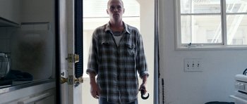 Movie still from “A Thousand Junkies” (2017), directed by Tommy Swerdlow – A man in a plaid shirt holding a wrench in a doorway; Medium shot, Low angle