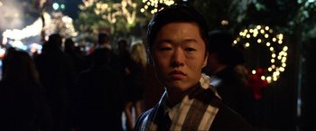 Movie still from “A Very Harold & Kumar Christmas” (2011), directed by Todd Strauss-Schulson – A young man wearing a scarf and a jacket is standing in front of a group of people; Close Up shot, Over the shoulder angle