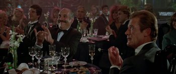 Movie still from “A View to a Kill” (1985), directed by John Glen – A group of people sitting at a table with wine glasses; Medium shot, Over the shoulder angle
