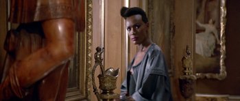 Movie still from “A View to a Kill” (1985), directed by John Glen – A woman in a gray shirt is standing next to a statue; Close Up shot, Low angle