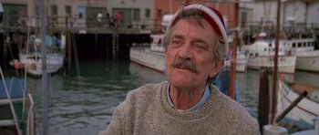 Movie still from “A View to a Kill” (1985), directed by John Glen – An older man wearing a sweater and a hat in front of a body of water; Close Up shot, Low angle