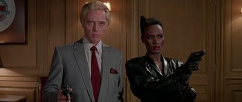 Movie still from “A View to a Kill” (1985), directed by John Glen – A man in a gray suit and a woman in a black leather jacket; Medium shot, Low angle