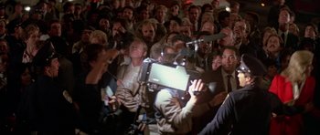 Movie still from “A View to a Kill” (1985), directed by John Glen – A group of people gathered around a man holding a camera; Wide shot, Over the shoulder angle