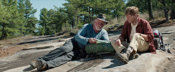 Movie still from “A Walk in the Woods” (2015), directed by Ken Kwapis – An older man sitting on the ground near a forest; Medium shot, Low angle