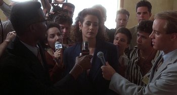 Movie still from “Ace Ventura: Pet Detective” (1994), directed by Tom Shadyac – A group of people gathered around a woman holding microphones; Close Up shot, Over the shoulder angle