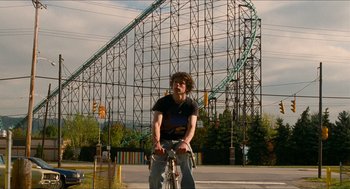 Movie still from “Adventureland” (2009), directed by Greg Mottola – A man riding a bike in front of a roller coaster; Wide shot, Low angle