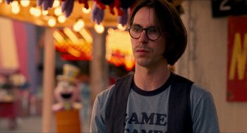 Movie still from “Adventureland” (2009), directed by Greg Mottola – A man wearing glasses and a t - shirt is standing in front of a carousel; Close Up shot, Over the shoulder angle