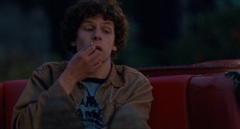 Movie still from “Adventureland” (2009), directed by Greg Mottola – A man sitting on top of a red bench smoking a cigarette; Medium shot, Over the shoulder angle