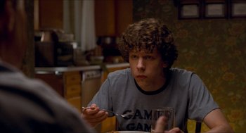 Movie still from “Adventureland” (2009), directed by Greg Mottola – A young man sitting at a dining table with a fork in his hand; Medium shot, Over the shoulder angle