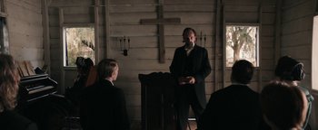 Movie still from “Alice” (2022), directed by Krystin Ver Linden – A man in a suit and tie standing in front of a cross; Medium shot, Over the shoulder angle