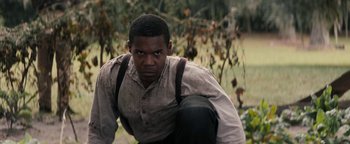 Movie still from “Alice” (2022), directed by Krystin Ver Linden – A black man in a white shirt and black pants crouching down; Medium shot, High angle