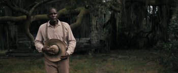 Movie still from “Alice” (2022), directed by Krystin Ver Linden – A man holding a hat standing in front of a tree; Medium shot, Low angle