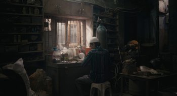 Movie still from “All That Breathes” (2022), directed by Shaunak Sen – A man sitting on top of a stool in front of a window; Wide shot, Low angle